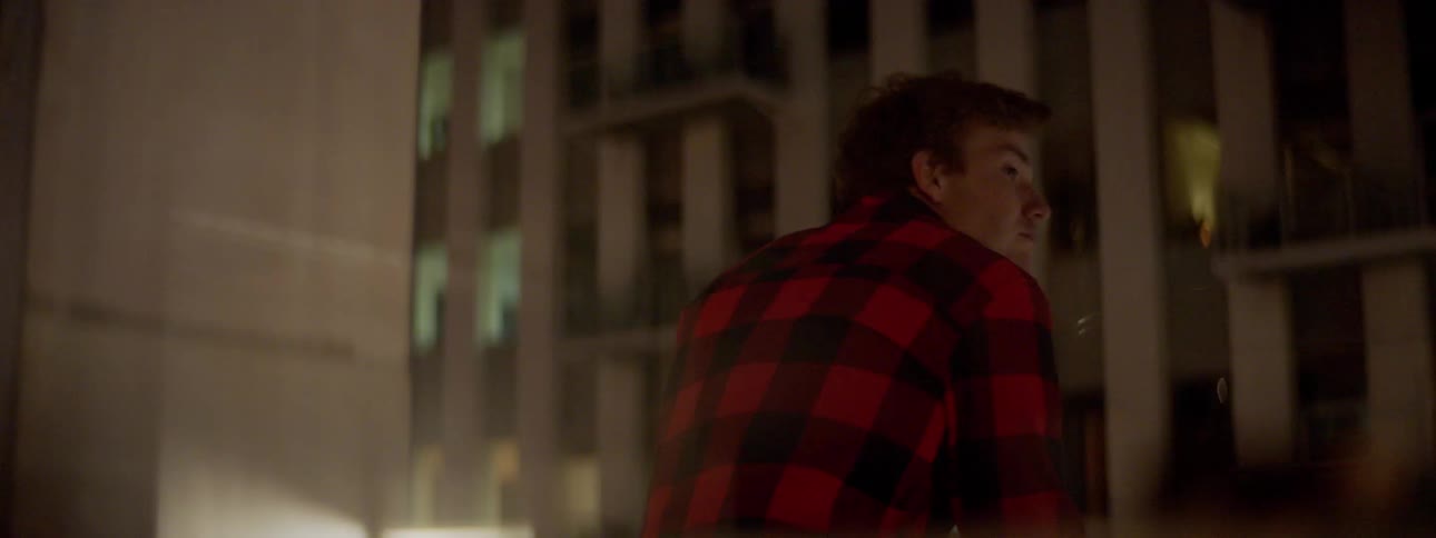 Man wearing a plaid shirt standing in the balcony of a building at night