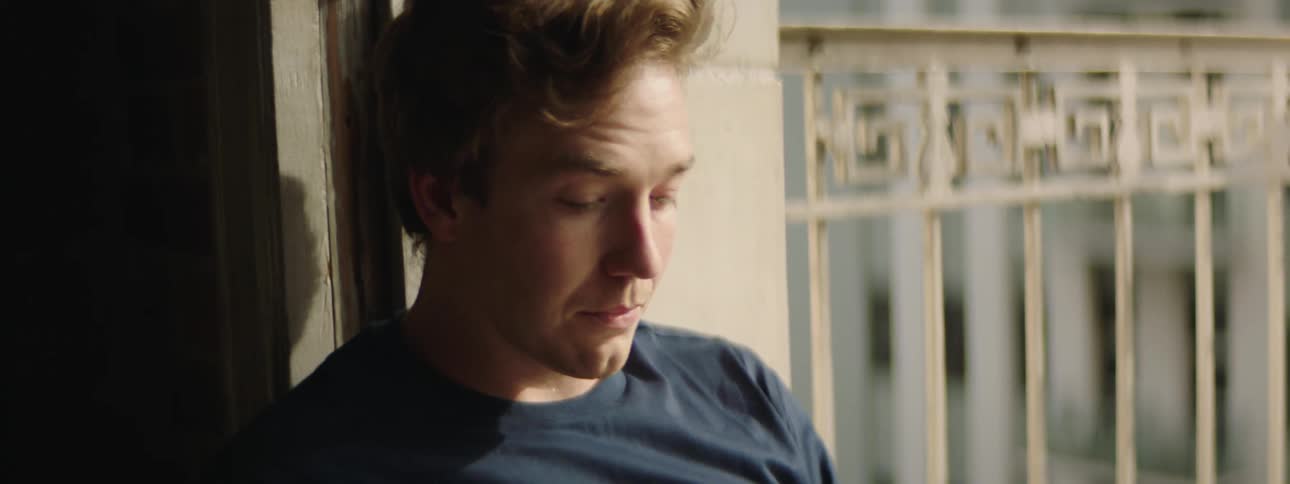 Man sits in sunlight by railing in blue t shirt looks sad rubs face and hand through hair