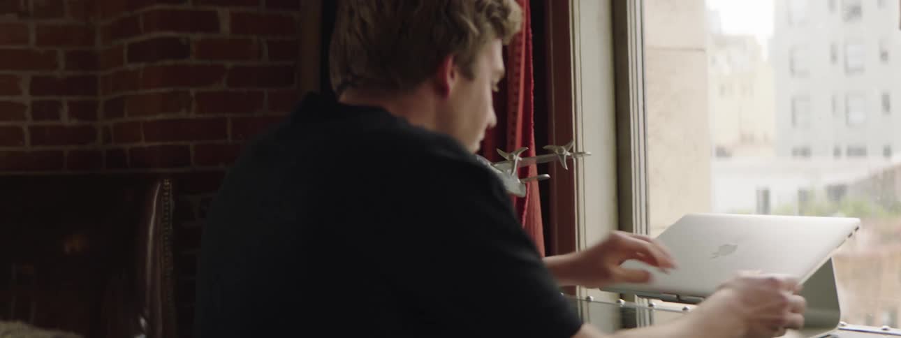 Man in black t shirt walks over to laptop on desk in room with brick walls and red curtains
