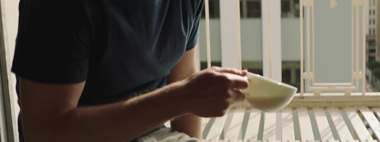 A man walks across his apartment picks up a mug from a table and walks over to sit on the balcony