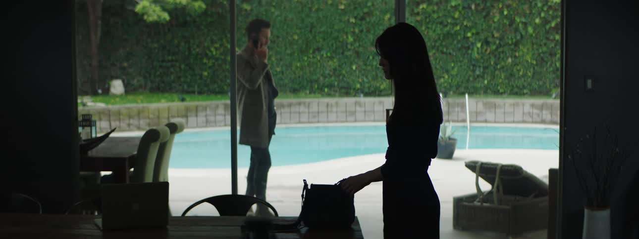 A man paces outside of a patio door while talking on a phone and a woman enters the room to get a purse