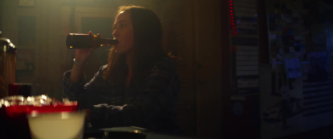 Woman in black jacket sits at bar in dark behind bright colored drinks drinking beer looks sad behind her is a door some posters and a string of lights