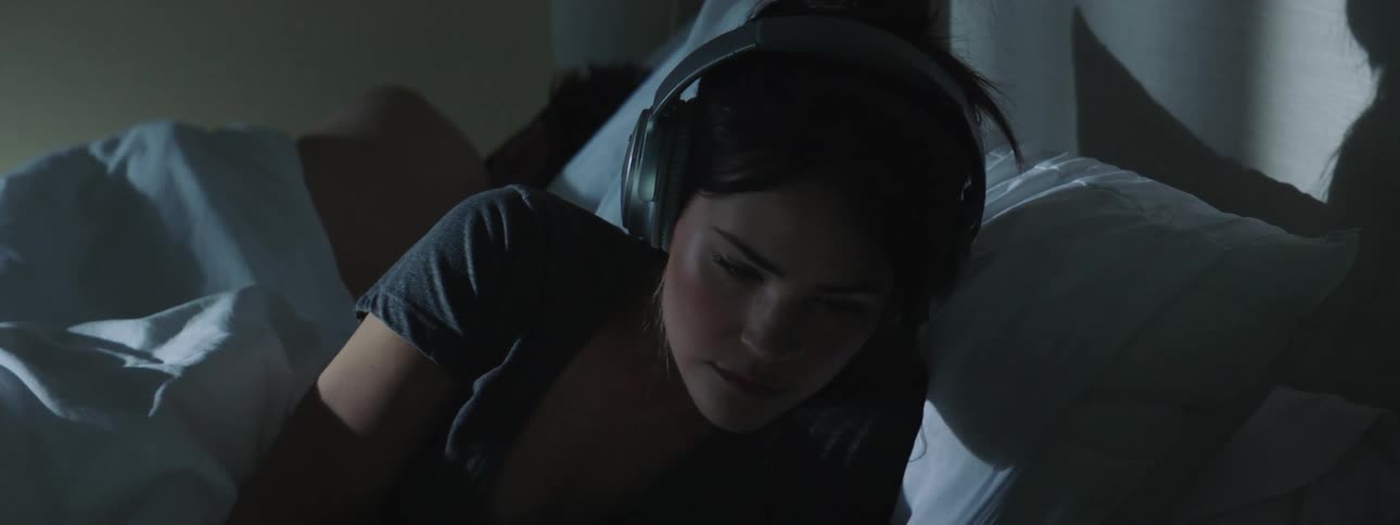 A woman lies in bed next to what appears to be someone else and wakes up picks up a pair of headphones and puts them on then closes her eyes to go back to sleep
