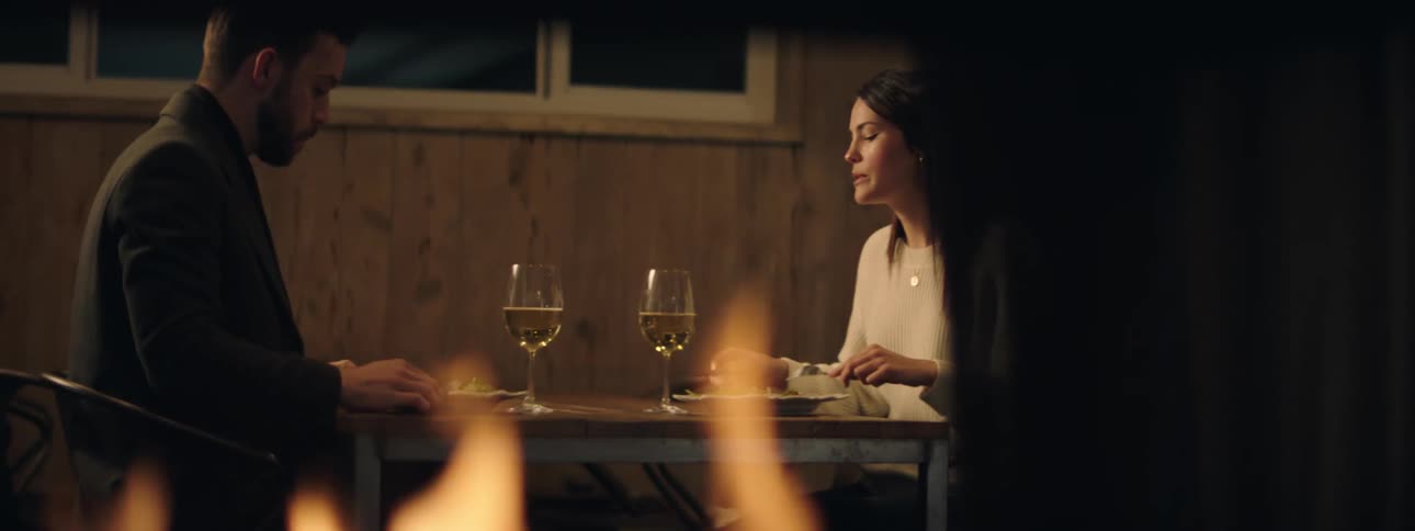 Man looks at his cell phone as he eats with a woman while a fire burns in a fireplace nearby