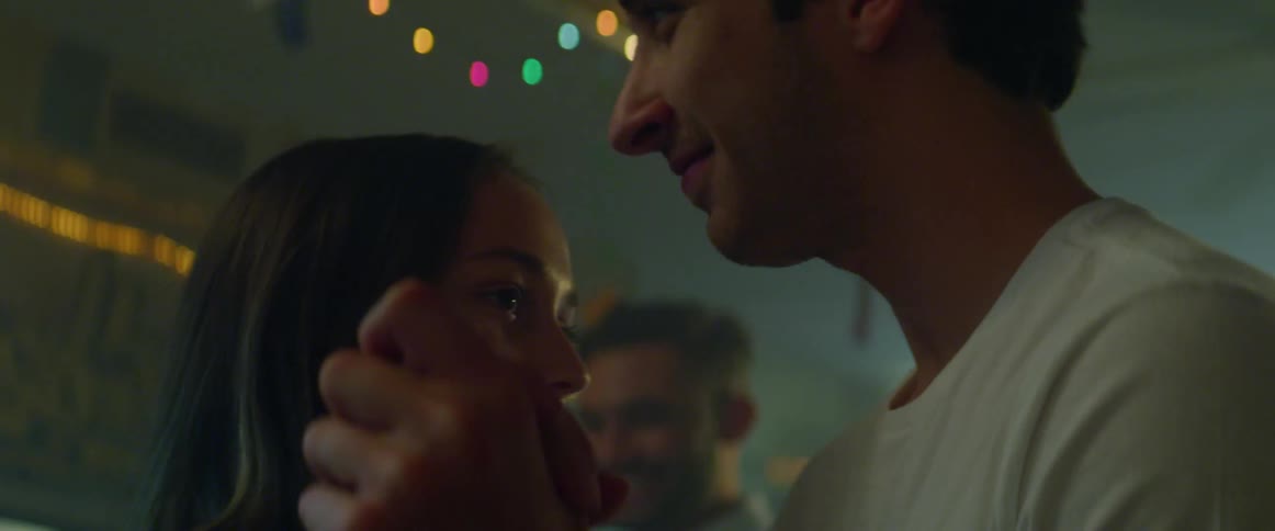 Man and a woman dance in a building decorated with hanging lights hand in hand with other people dancing behind them and the sun showing through the door