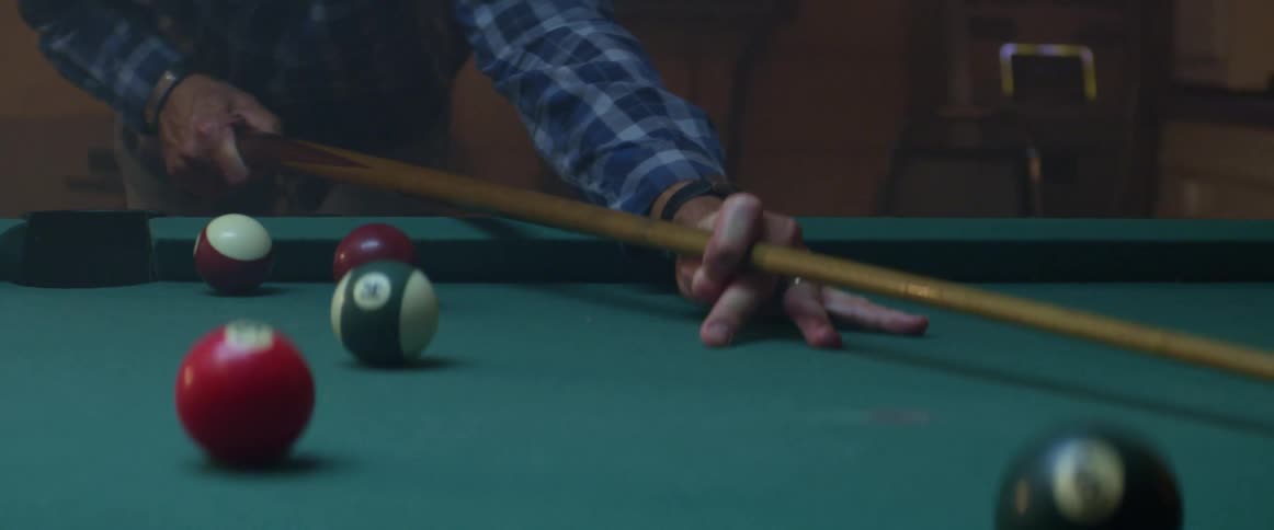 A man in a blue flannel shirt plays a game of pool alone