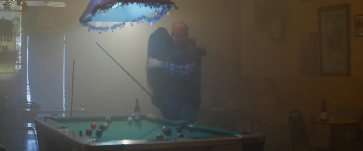 A woman puts a beer down on a pool table and hugs a man holding a pool cue