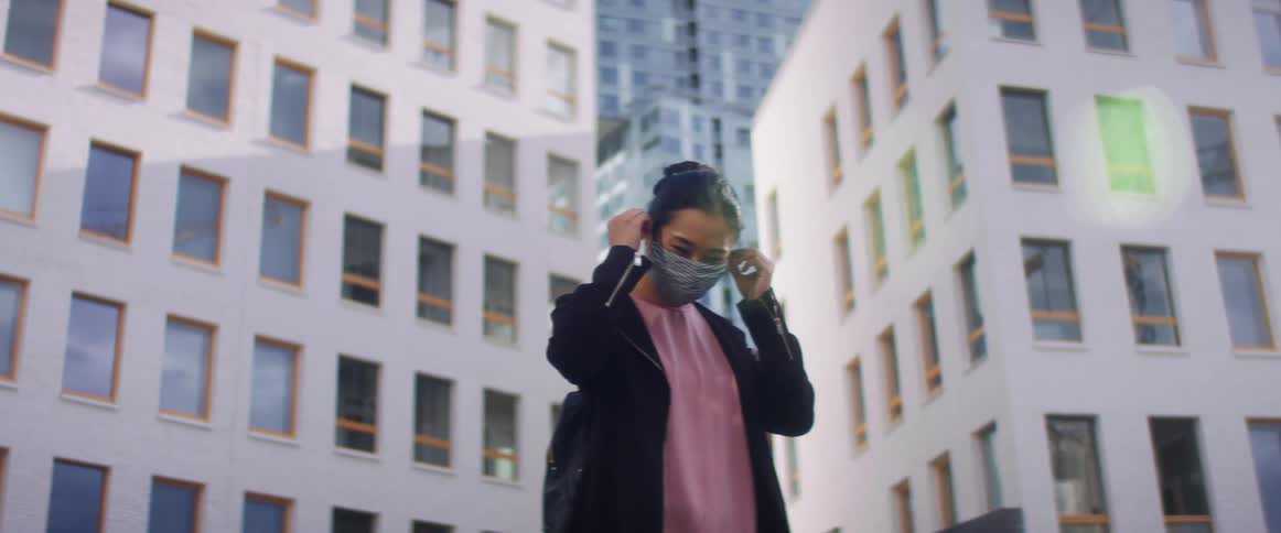 A woman stands in front of large city buildings and places a face mask over her mouth and nose