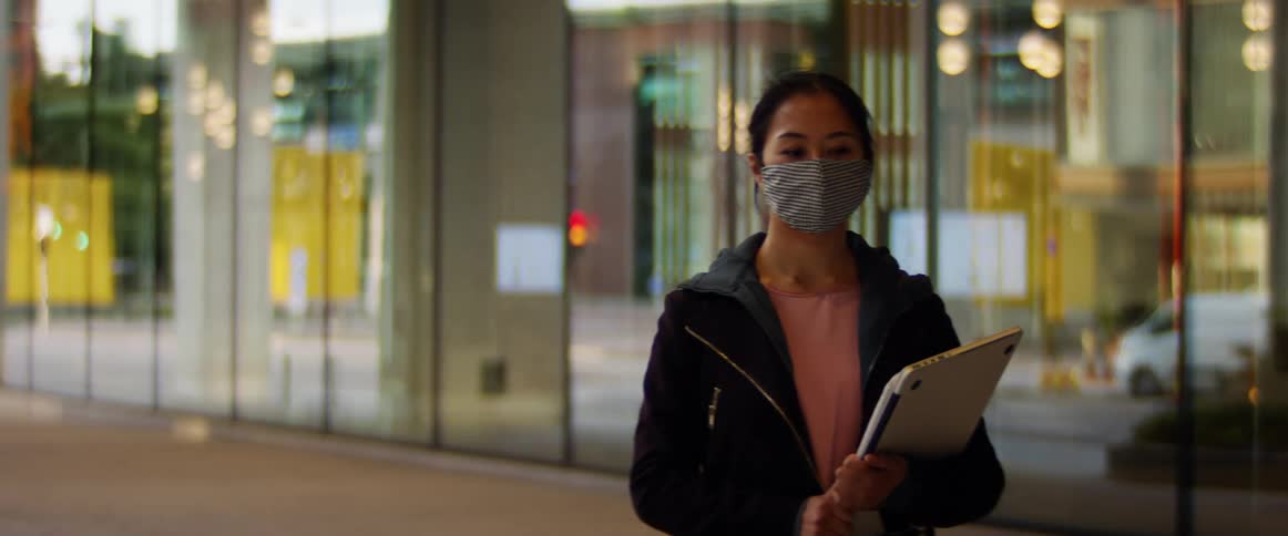 A woman wearing a face mask carries a clipboard as she walks on a sidewalk in front of a building