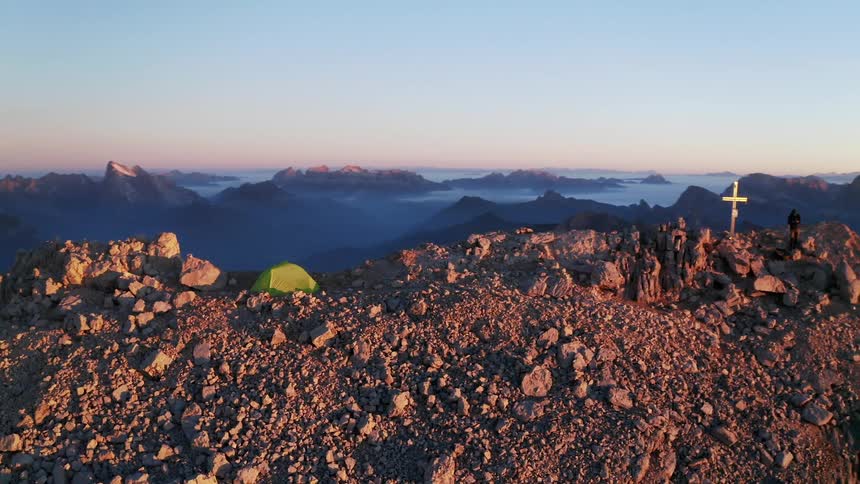 A person camping on the summit of a mountain stands near a cross erected on the peak
