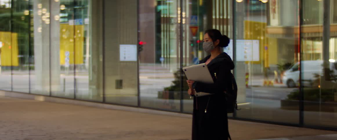 A woman carrying a laptop walks in front of an office with vehicle reflecting in the windows