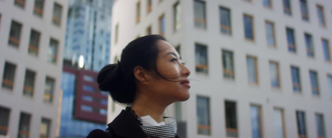 Woman standing outside near large buildings looking up and ahead of her with the wind blowing in her hair