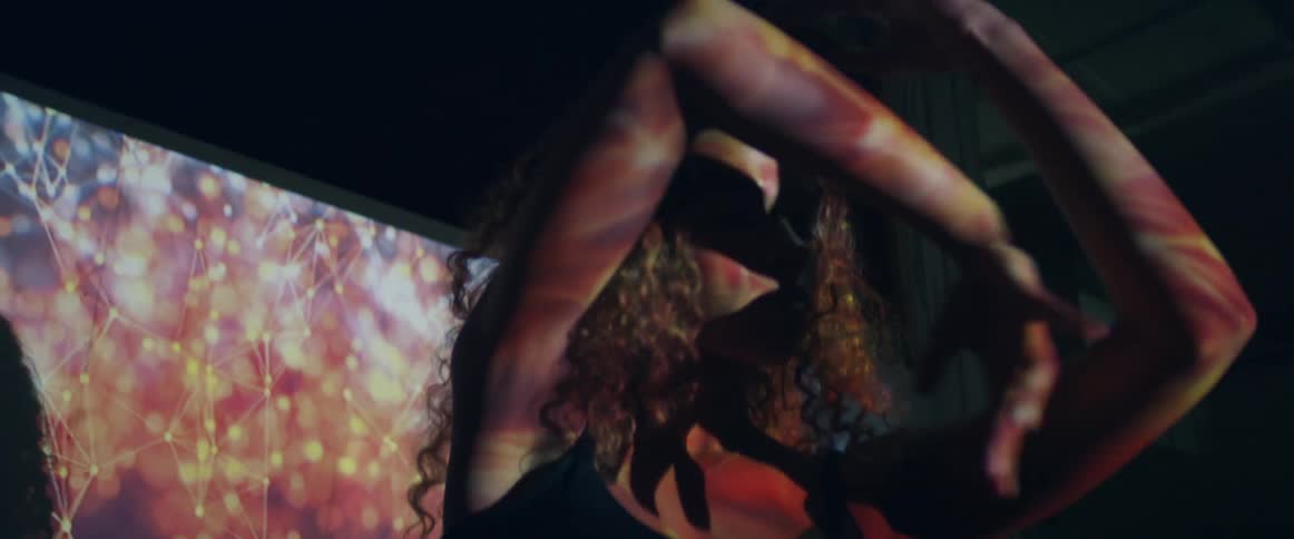 A woman dances slowly moving her arms on a stage in front of a screen with colors of light