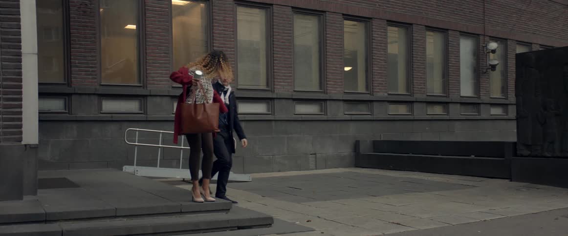 A woman stands outside of a building and struggles to put on a red coat when a man exits the building and helps her put on the jacket by lifting the arm sleeve to her hand