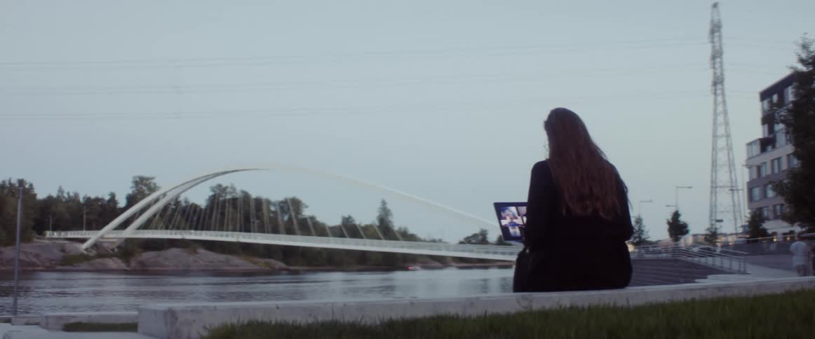 A woman sits in front of the water in a city and looks at her laptop as pedestrians walk past