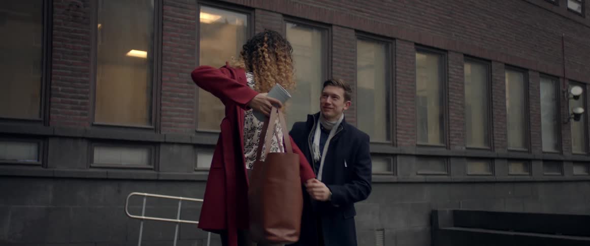 A woman standing in front on an office building is struggling to put her jacket on while holding a coffee cup and tote bag when a man approaches from behind and helps her