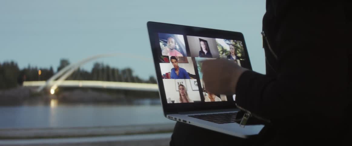 A woman is sitting indoor by window with laptop on her lap doing a zoom meeting with other people