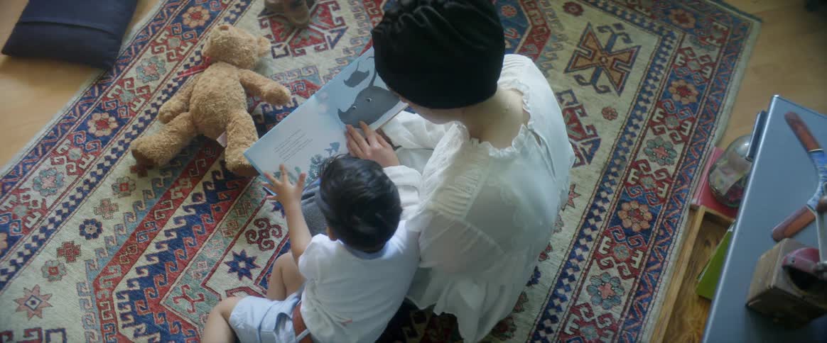 A woman sitting on a rug while reading a picture book with her young son