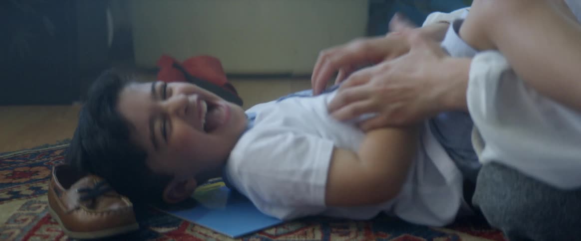 A little boy lies on the floor at home and laughs as his mother tickles him