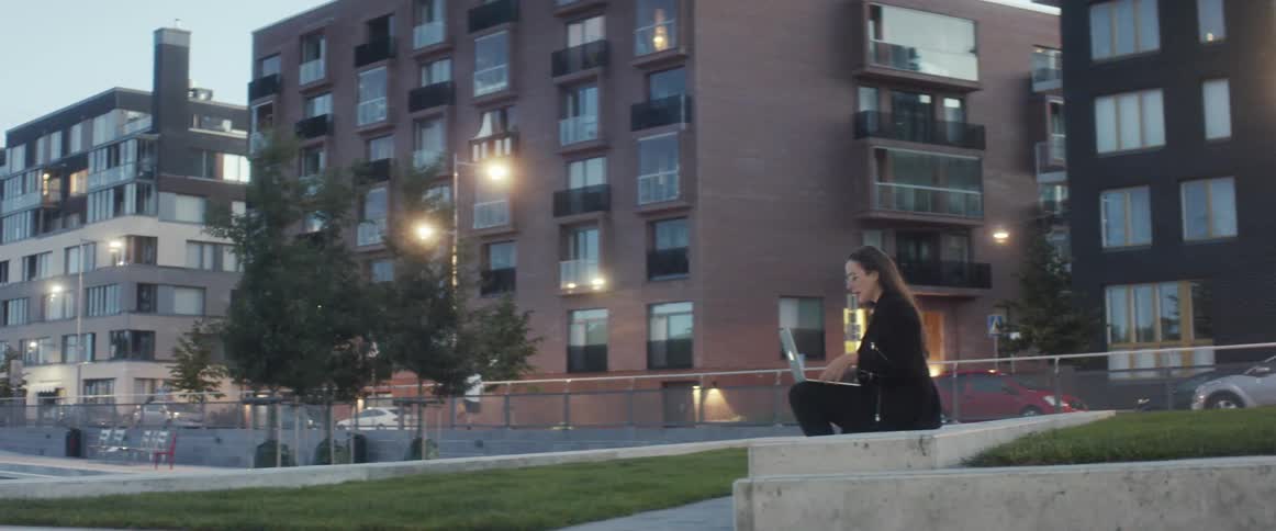 Woman in black clothing wearing glasses sitting on concrete in a park a lap top computer on her lap speaking to someone via face timing a pedestrian across the street walking as a car on the street passes by