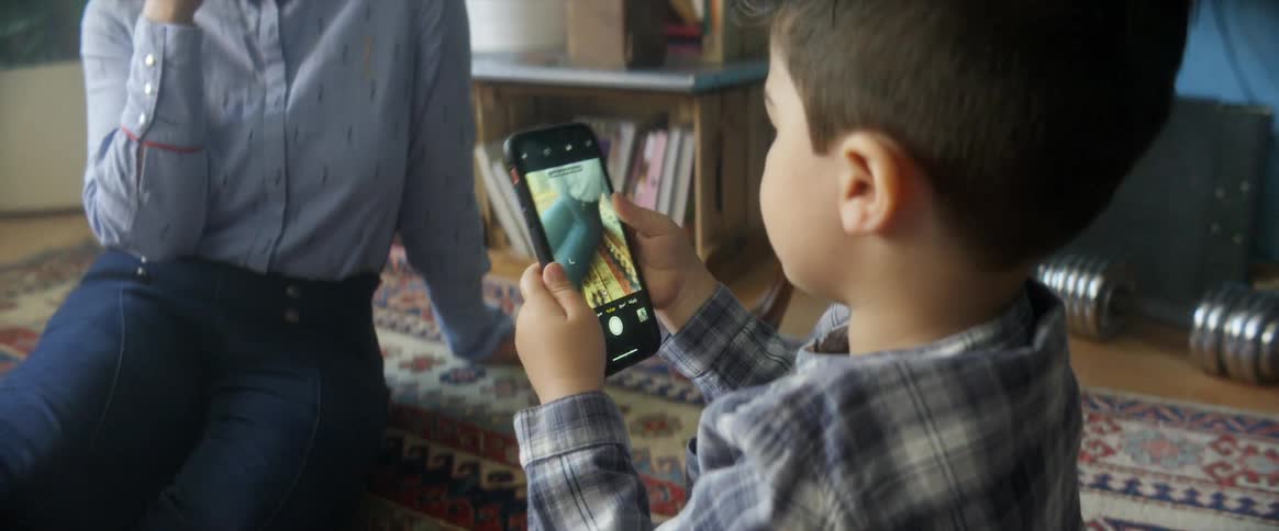 Mother sits in the floor on a rug and poses as son takes pictures on a smart phone there are plants books a weight a internet sever and other household items behind them