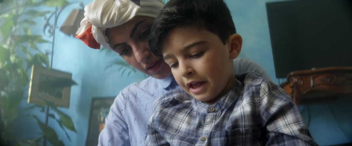 Mother holds son in lap while they look at the cellphone he is holding while sitting in a lit room in front of a blue wall and television on a table