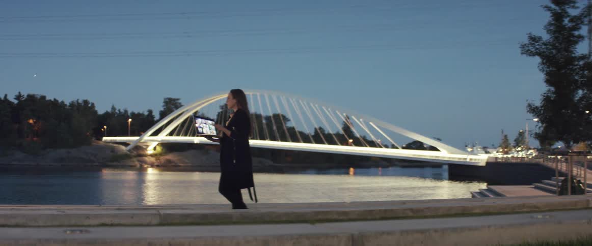 A woman standing on the shore of a river in the evening participates in a video chat using the laptop she holds with one arm