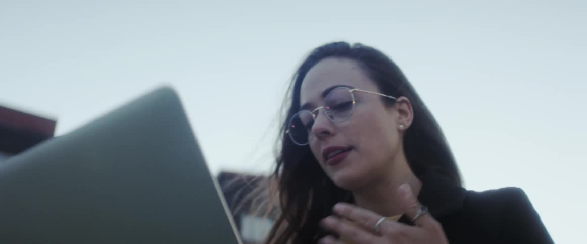 A woman is communicating with her laptop as she speaks as she holds the laptop in her hands outside a building in windy cold looking weather