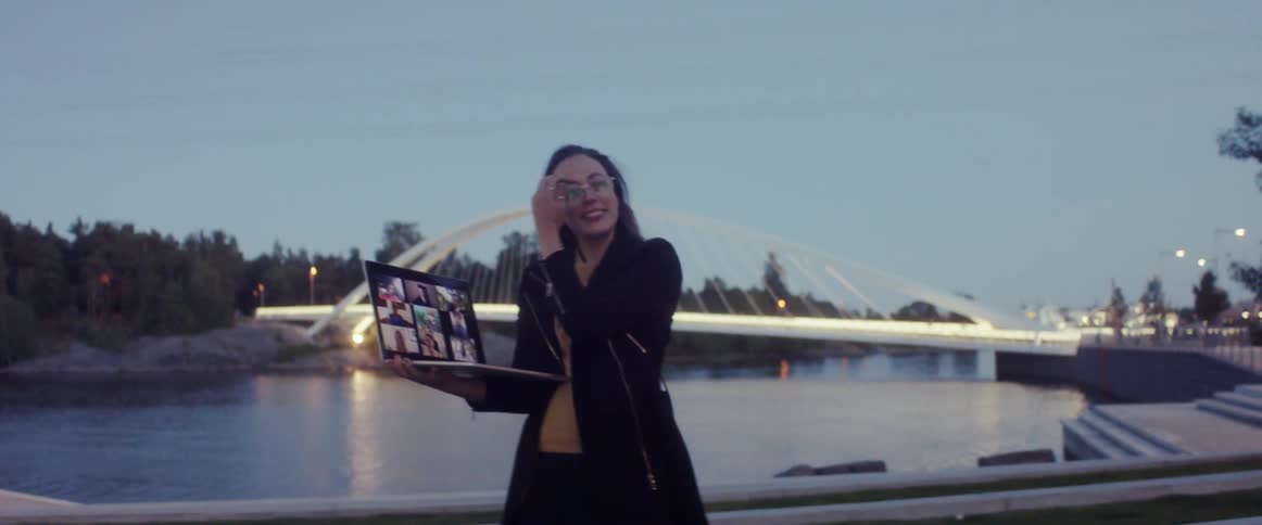 A woman shows a group of people her city via video call by holding her laptop while standing in front of a reservoir in a city