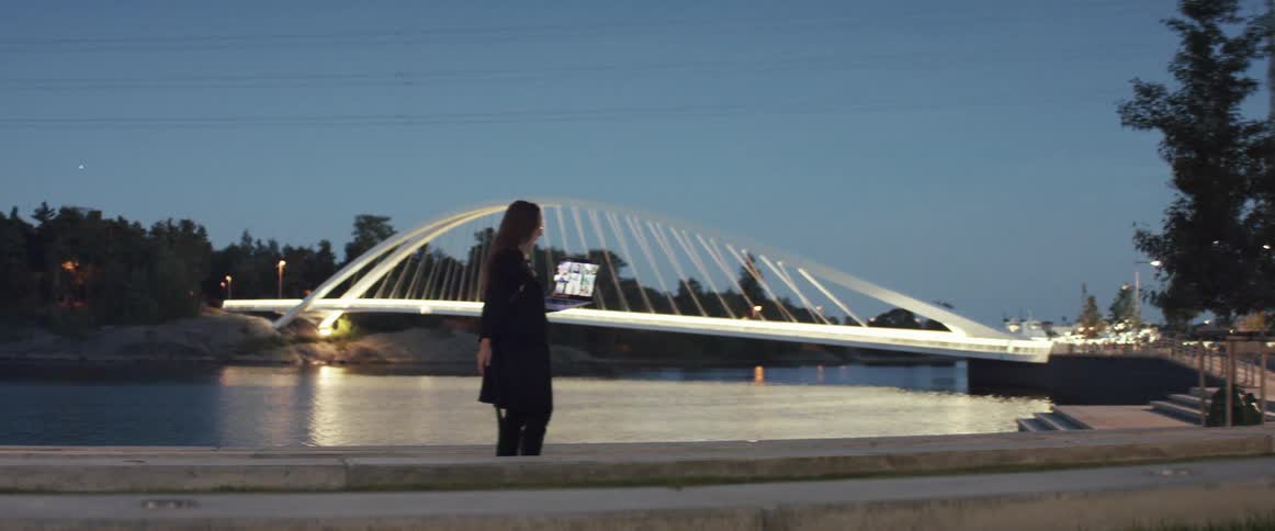 A woman stands in a park in front of the water and talks as she hold her laptop and looks at it
