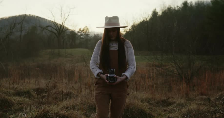 A woman with long hair and a hat looks down at a camera in her hand while standing outdoors in nature on an overcast fall day