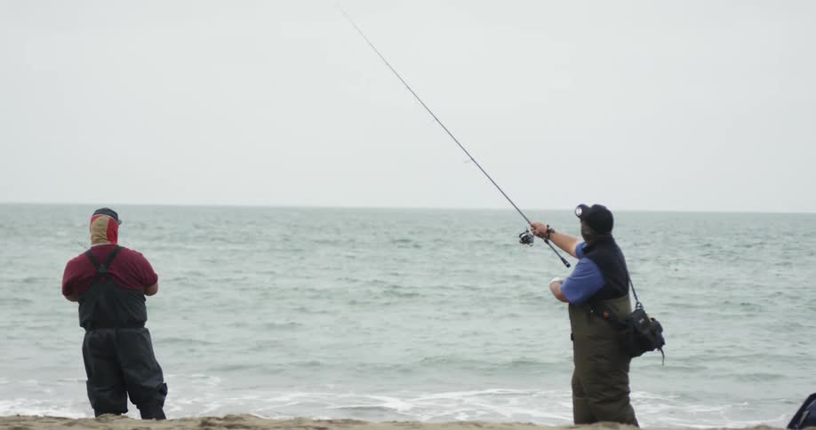 Two men in fishing gear cast fishing lines out into the ocean right off the shore
