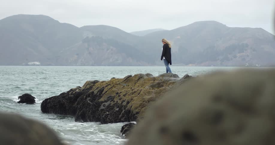 Woman is walking on large rock on the ocean as wind blows and waves splash