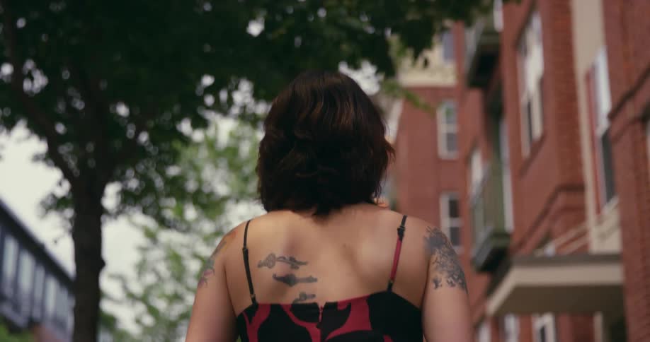 Woman with tattoos walks along sidewalk past apartment buildings