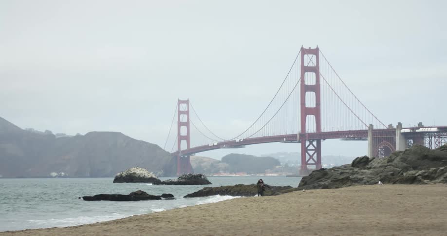 Waves crash on beach under bridge around rocks with cliffs in the distance on a misty foggy cloudy day