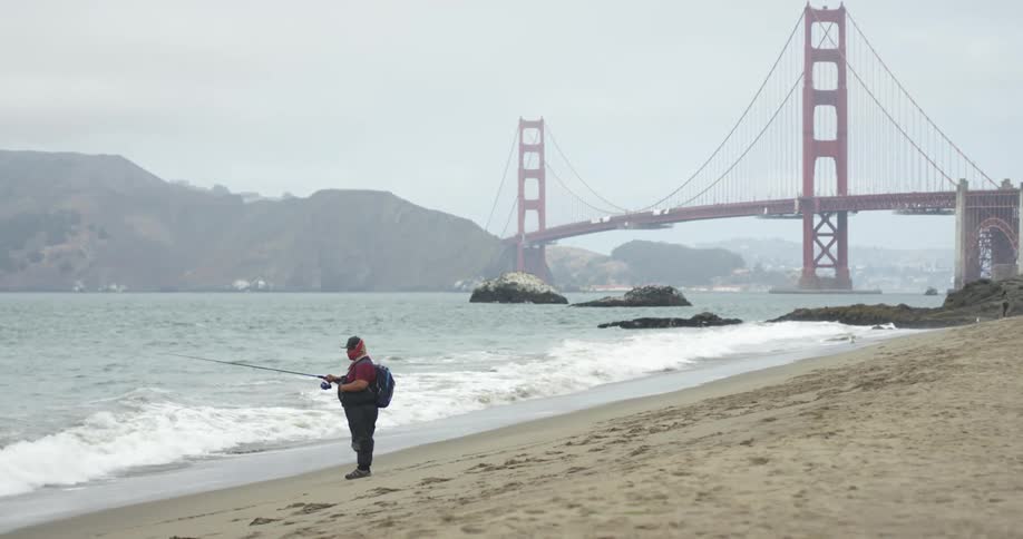 A man with a hat and a backpack fishing on a beach with a bridge the sky and clouds in the background