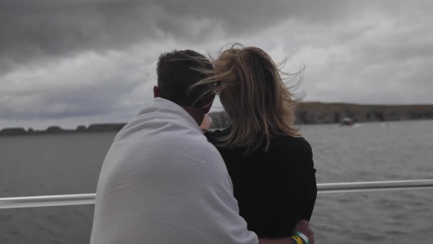 Man stands on boat holding a woman who points out over the water a rocky shore line in the distance under a grey cloudy sky