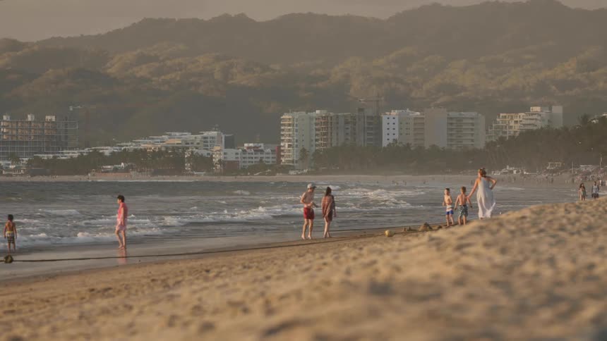 A large group of people are on a beach with crashing waves walking around as a child plays and runs around with his family