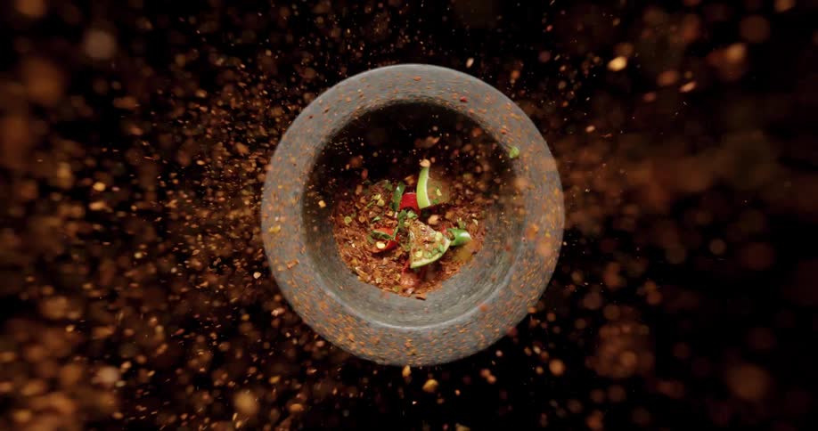Red spices are sprinkled on limes and peppers in a stone bowl that is spinning with a back background