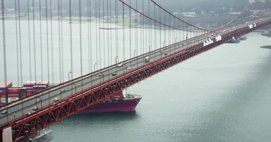 A boat in a large body of water drives by under a giant red bridge where there are cars driving on the bridge