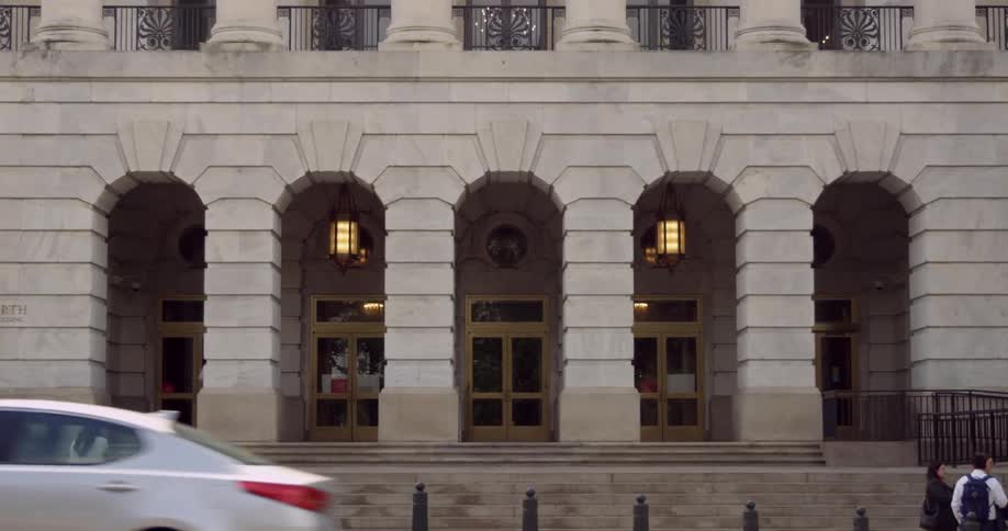 People walk into a large downtown building with arched entrances as traffic passes by