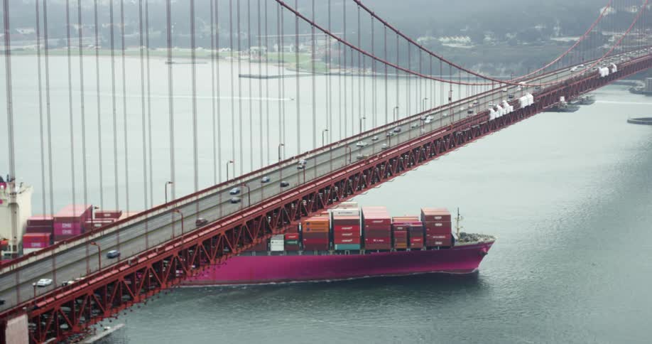A boat carrying shipping containers sails under a bridge with cars driving over it