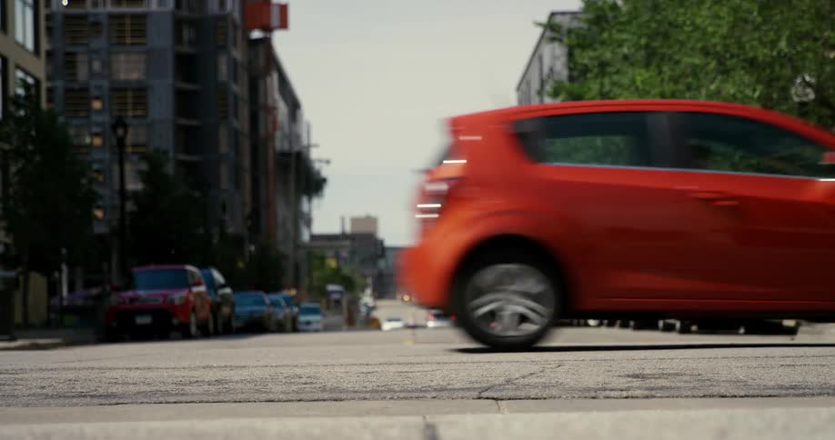 Red car passes on city street with parked cars and cars stopped at red lights with trees blowing in the wind beside buildings