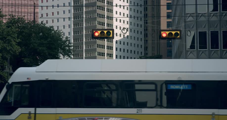 Trains are moving past a traffic light in the middle of a city with traffic around and building in the background