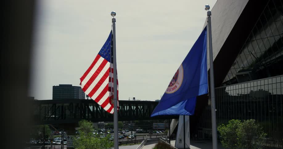 Two flags outside on flagpoles flutter in the wind above a full parking lot and trees and beside and in front of buildings