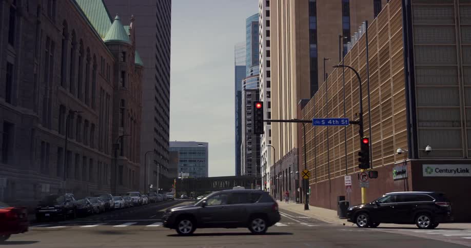 A city street with traffic traffic lights and a person crossing at the crosswalk with tall buildings going down and cars parked down the sides on a sunny day