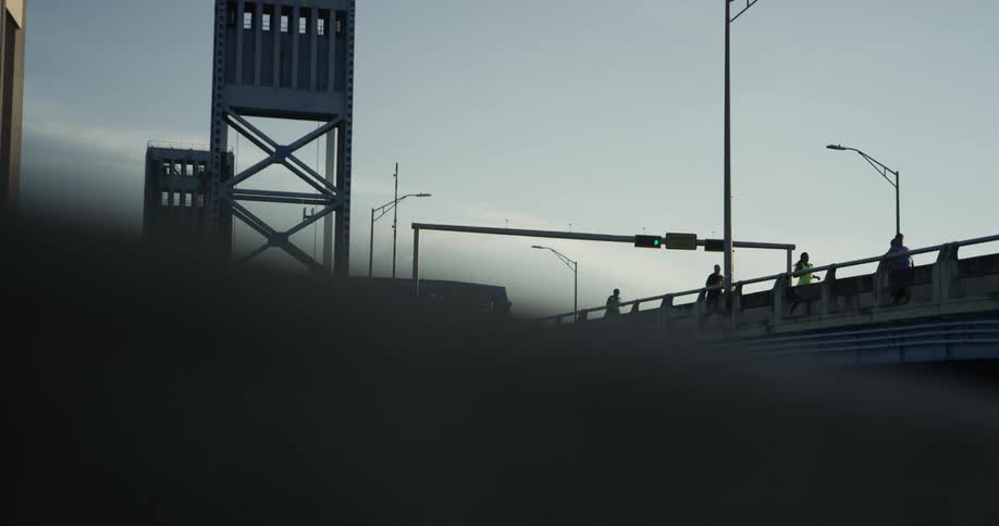 People are walking and jogging on a bridge with little sunlight and a traffic and street light present