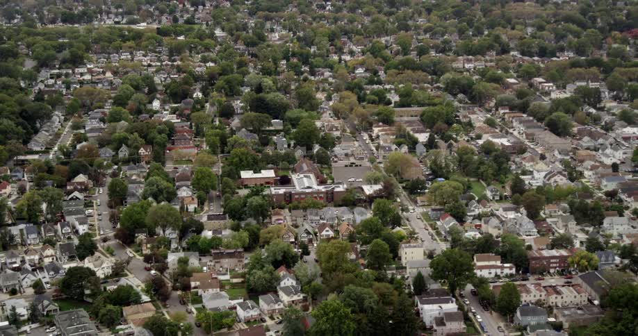 A camera hovers over a large neighborhood full of houses and trees with other recreational areas about and parked vehicles