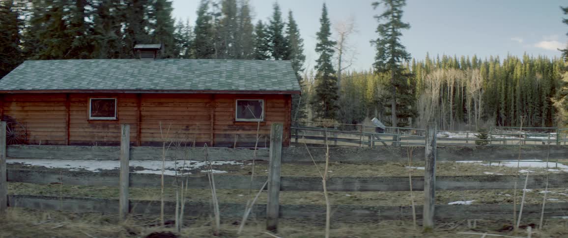 A house out in a rural setting displayed in front of a forest where there is snow on the ground surrounded by a wooden house
