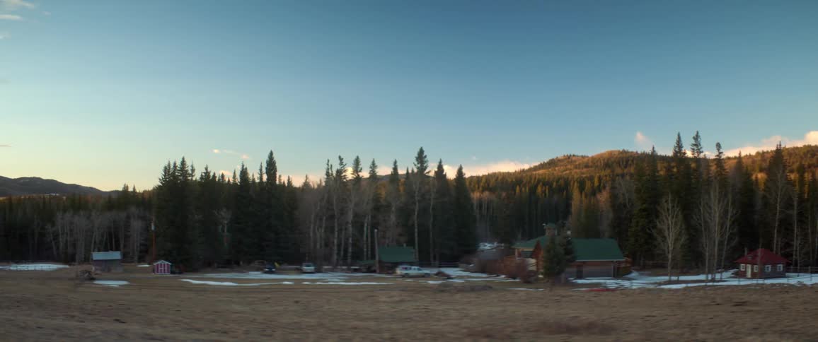 A house and a small structure are against a line of trees and hills in the background with a low sun and a little snow on the ground